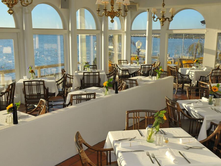 A restaurant with white tables and chairs, large windows with a view of the sea, and a chandelier.