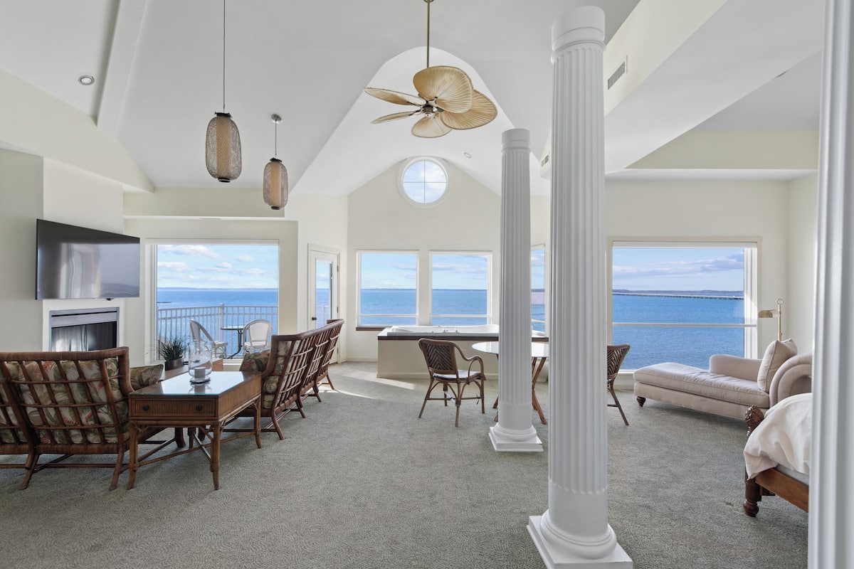 A spacious living area with a dining table, chairs, a sofa, and a large window overlooking the sea.