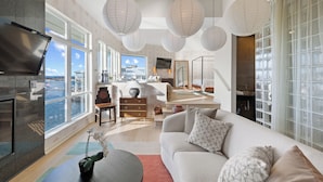 A modern living room with a large sofa, a coffee table, and a TV. There are large windows with a view of the water and a balcony.