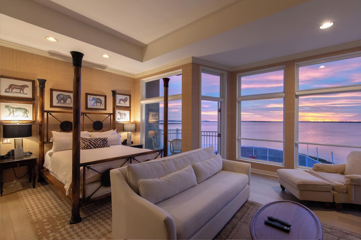 A spacious room with a large bed, a sofa, and a balcony with a view of the sunset over the water.