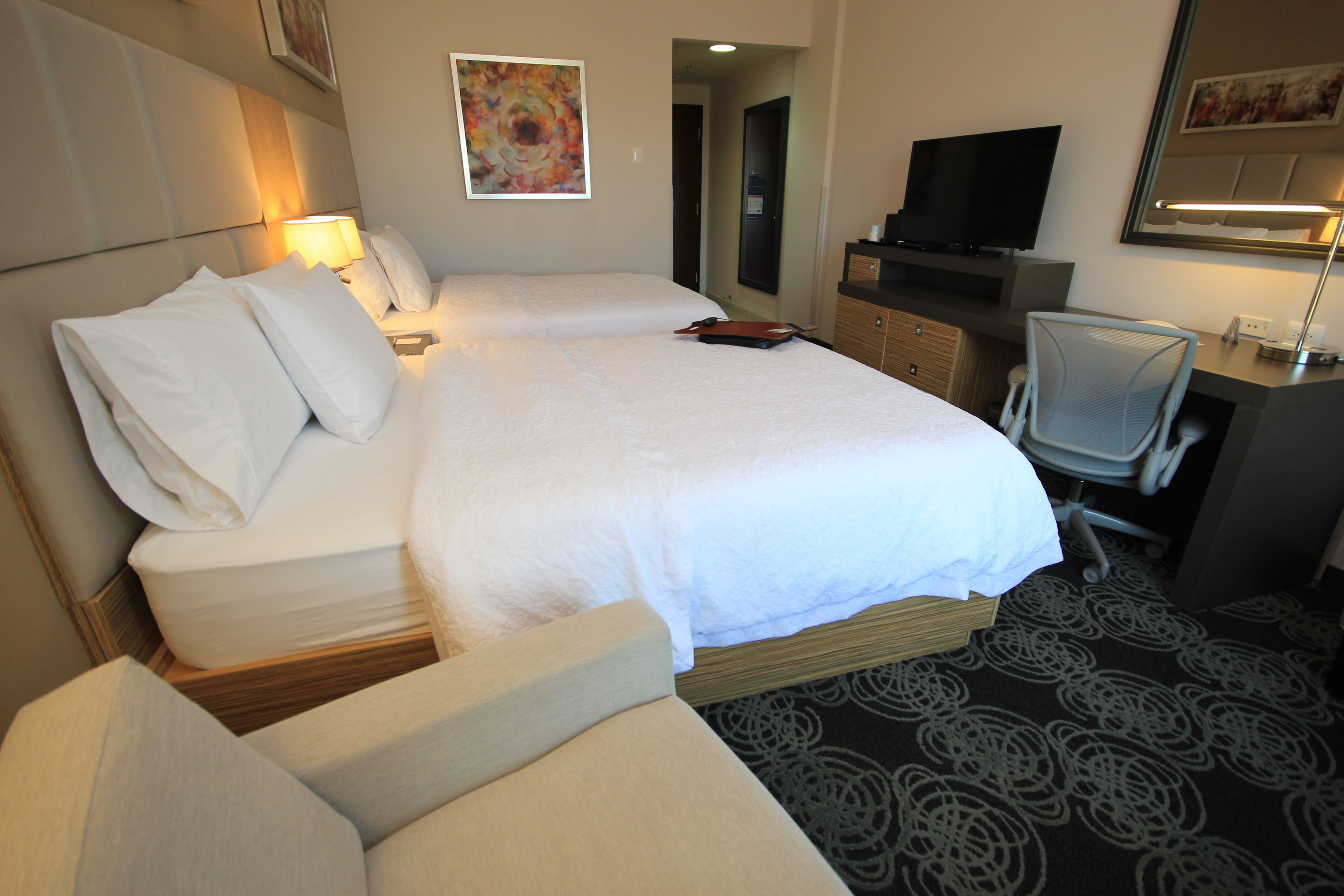 Ein Hotelzimmer mit einem großen Bett, einem Schreibtisch mit Stuhl, einem Fernseher und Bildern an den Wänden.