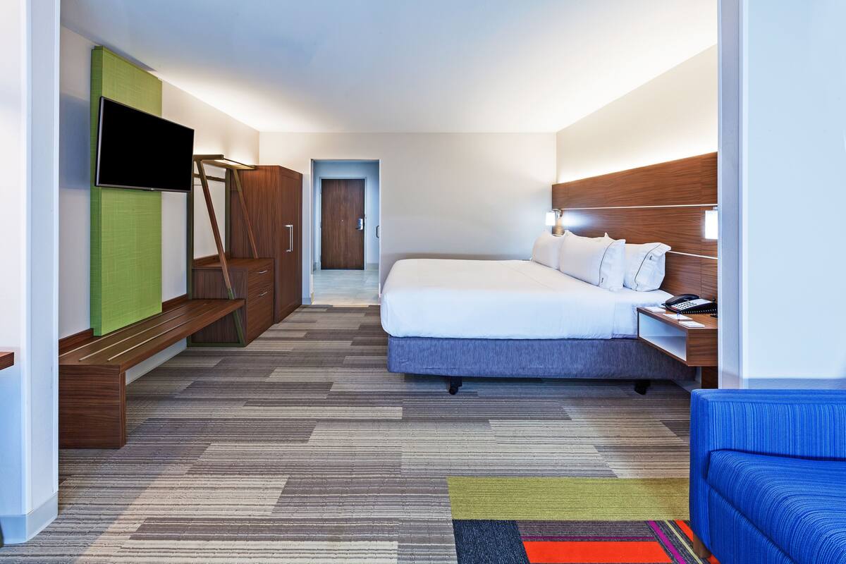 A hotel room with a bed, a TV, a desk with a lamp, a bench, and a wardrobe.