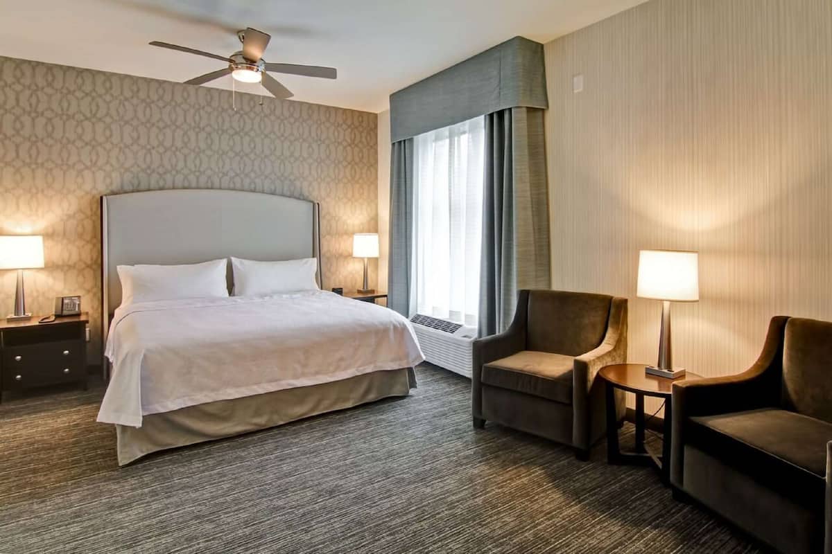 Une chambre d’hôtel avec un grand lit, deux fauteuils, une petite table, un ventilateur de plafond et une grande fenêtre avec des rideaux.
