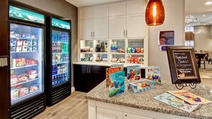 A hotel lobby with a kitchenette area, including refrigerators stocked with food and beverages, a coffee machine, and a chalkboard sign. - Homewood Suites by Hilton Clifton Park (Clifton Park)
