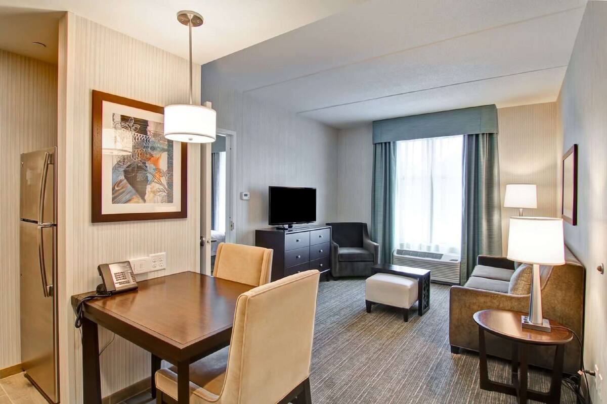 Une chambre d’hôtel comprenant un coin repas, un espace salon avec un téléviseur et une grande fenêtre avec des rideaux.
