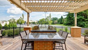 An outdoor patio with a stone table and chairs, a pergola with string lights, and a view of a parking lot and trees. - Homewood Suites by Hilton Clifton Park (Clifton Park)