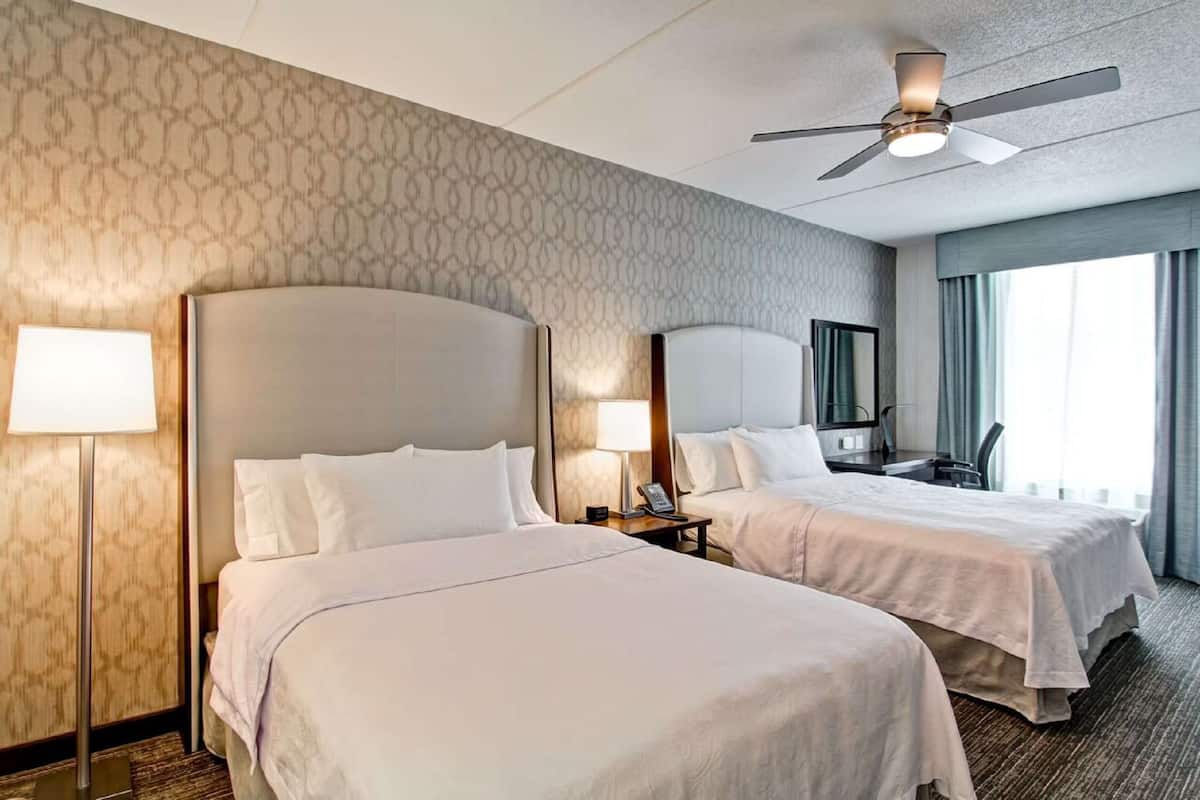 Une chambre d’hôtel avec deux lits, un ventilateur de plafond, un bureau avec une chaise et une grande fenêtre avec des rideaux.