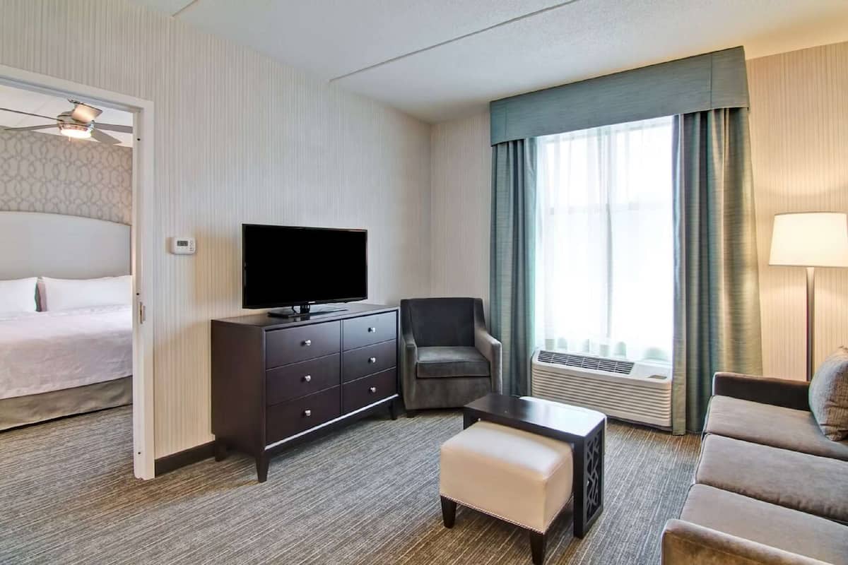 Une chambre d’hôtel avec un lit, un téléviseur posé sur une commode, une chaise, un canapé et une fenêtre avec des rideaux.