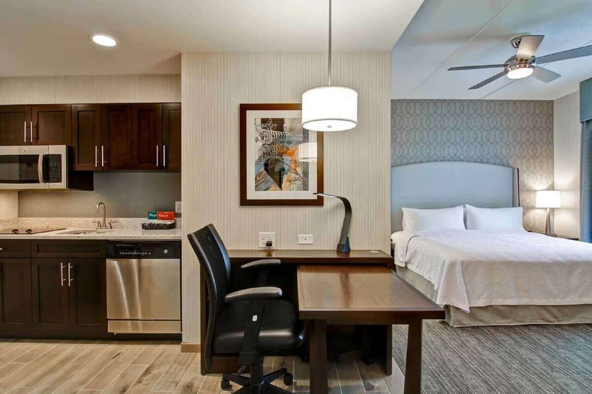 Ein Hotelzimmer mit Bett, Schreibtisch, Stuhl, Küchenzeile und Deckenventilator.
