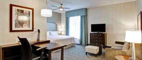 Ein Hotelzimmer mit Bett, Schreibtisch, Bürostuhl, Sessel, Fernseher und Deckenventilator.