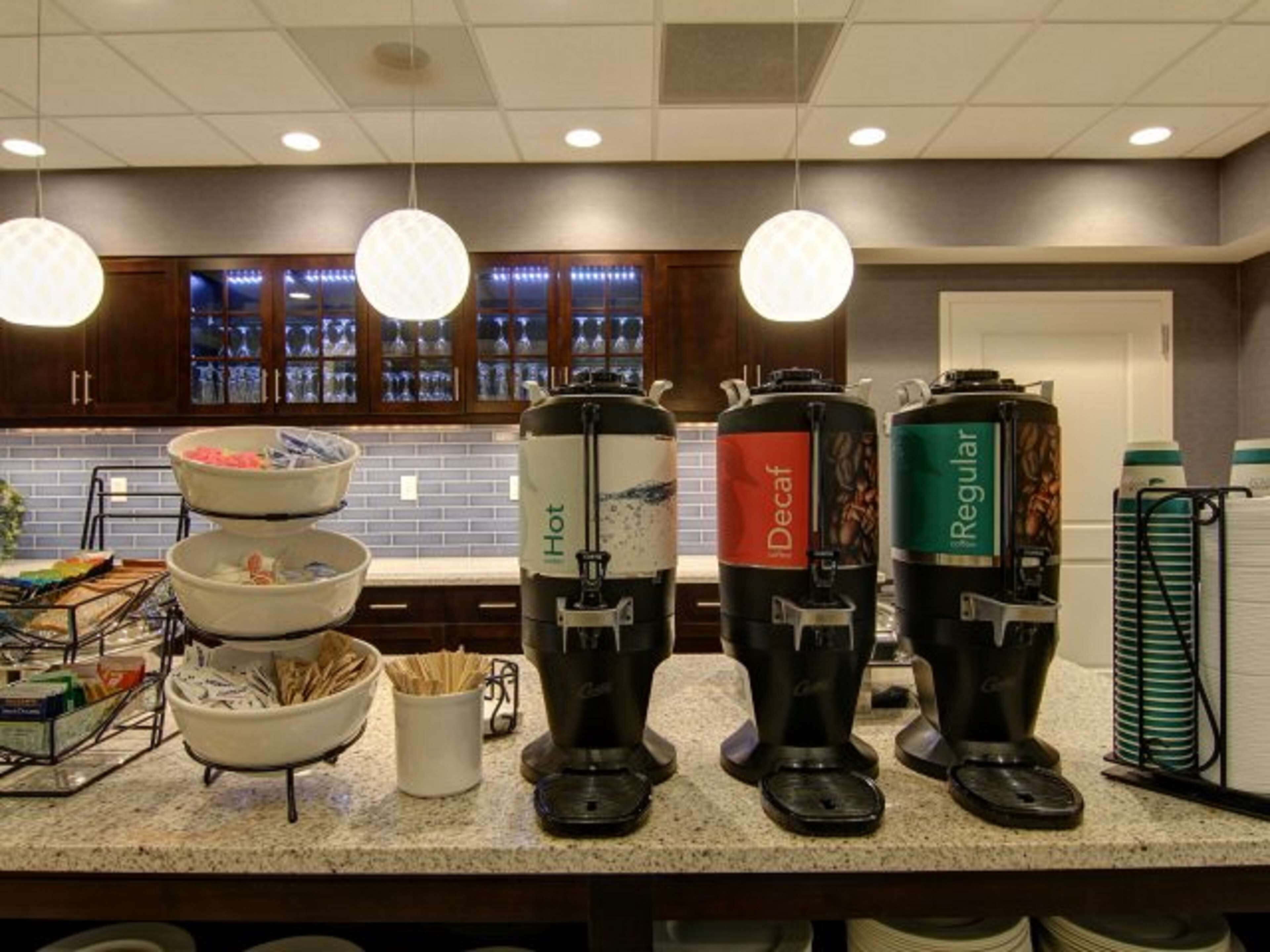Una estación de café con tres dispensadores de café etiquetados 'Hot', 'Descafeinado' y 'Regular'. Hay cuencos con bocadillos y una cafetera sobre el mostrador.