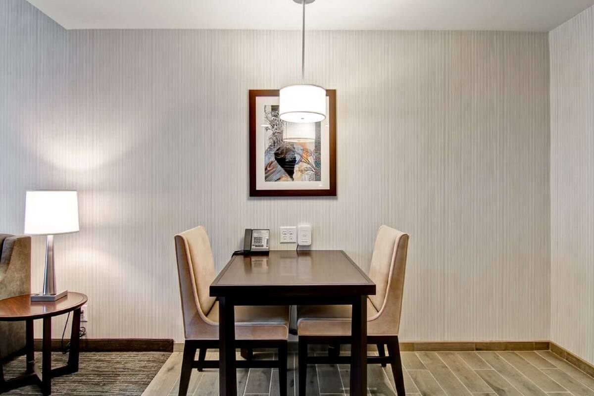 Une chambre d’hôtel dotée d’un coin repas comprenant une table en bois et deux chaises, une œuvre d’art encadrée au mur et un téléphone sur la table.