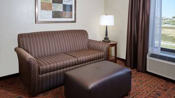 Una camera d'albergo con un divano a righe, un tavolino con una lampada, una moquette fantasia e una finestra con tende.