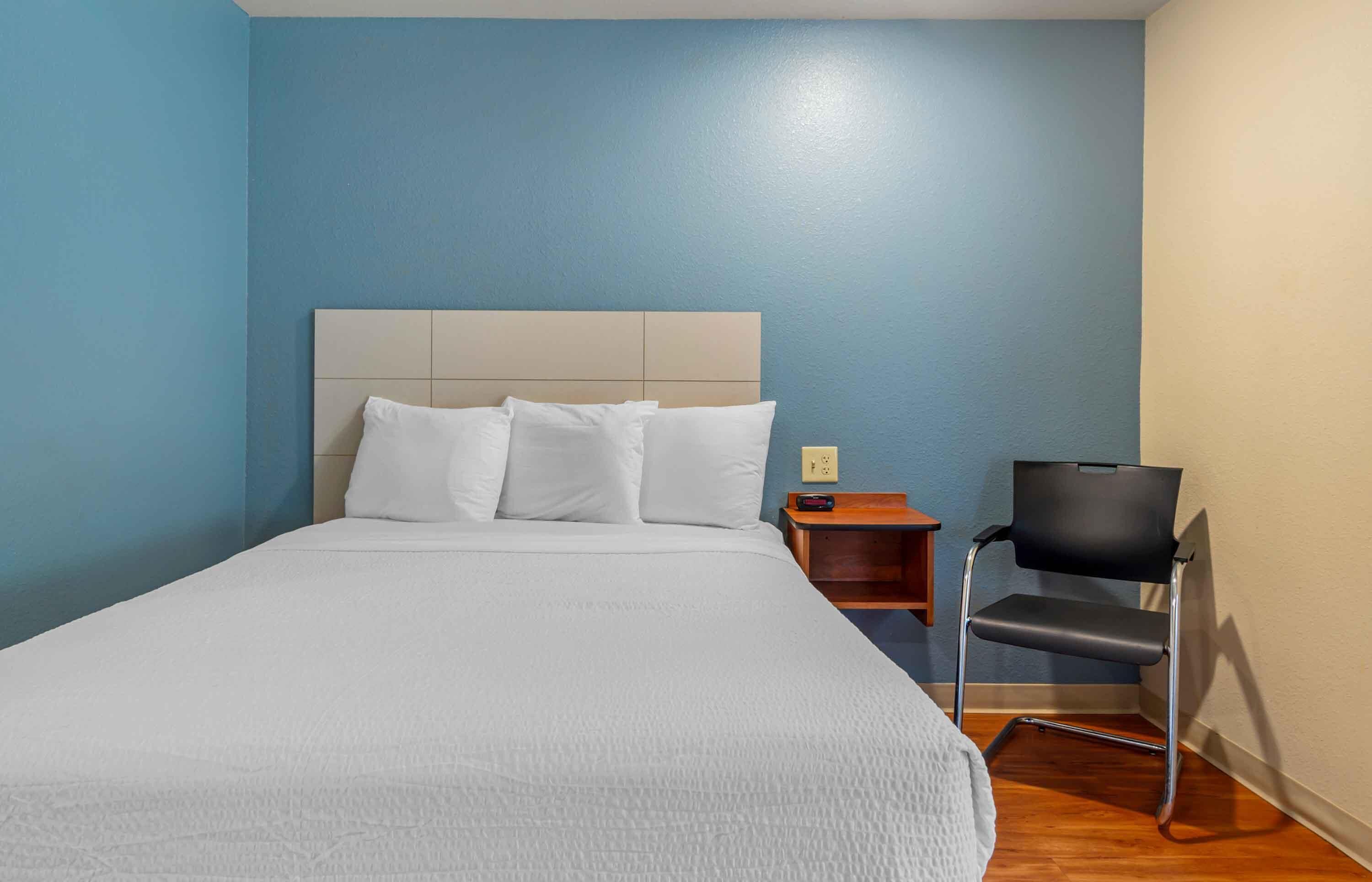 A hotel room with a large bed, a wooden nightstand, a modern chair, and a blue wall.