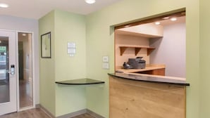 A reception area with a wooden counter, a glass partition, and a door leading to a hallway. - Extended Stay America Select Suites - Atlanta - Chamblee (Chamblee)