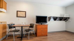A compact room with a wooden dining table, two chairs, a TV on a stand, a small cabinet, and a wall-mounted shelf with a basket. - Extended Stay America Select Suites - Atlanta - Chamblee (Chamblee)