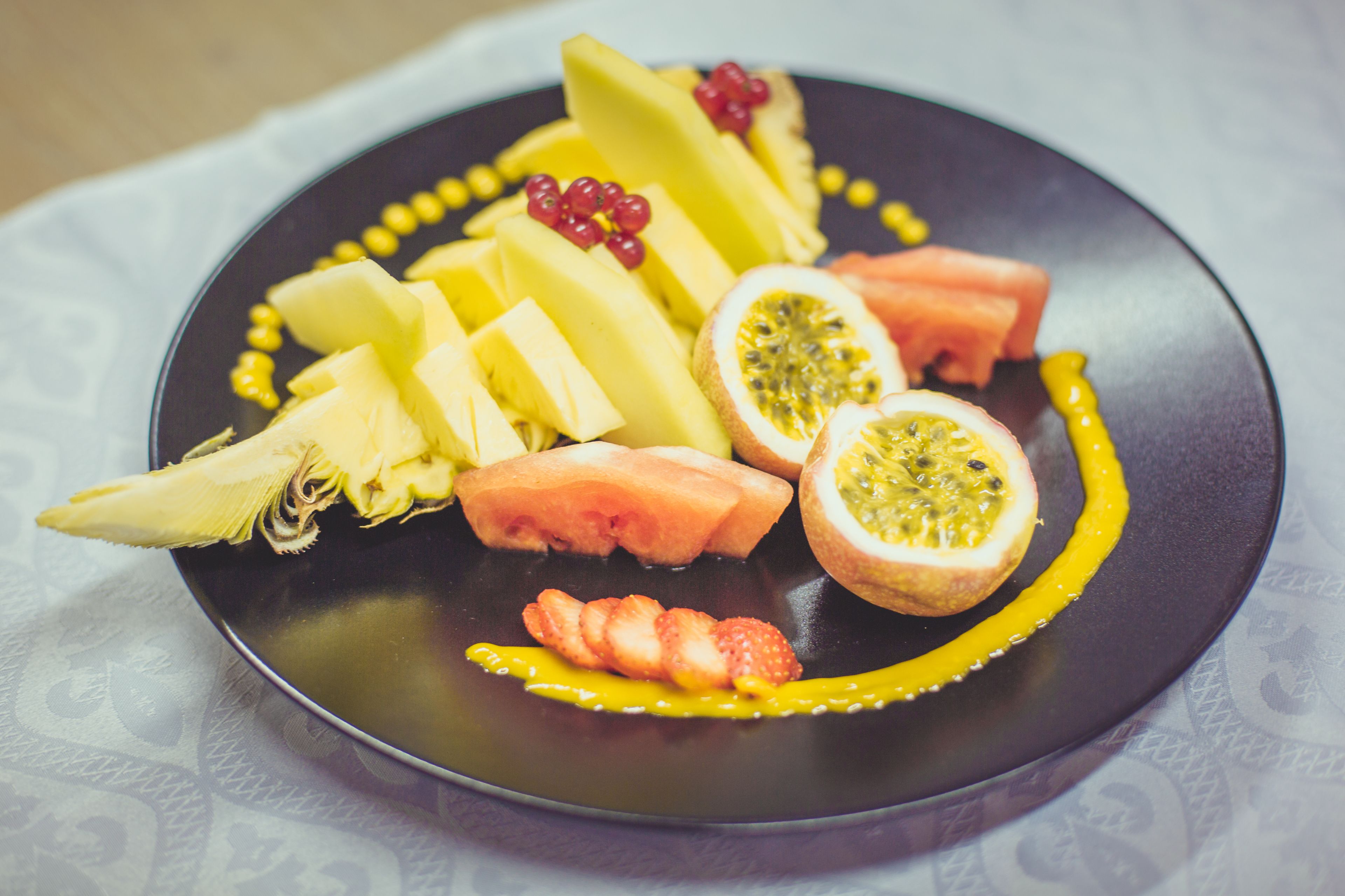 Prato com fatias de frutas como melancia, kiwi e manga, disposto sobre um prato escuro com um fio de calda amarela.