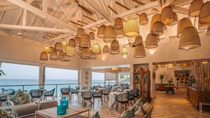 Een ruime eetruimte met meerdere tafels en stoelen, grote ramen met uitzicht op zee en hangende rieten lampen.