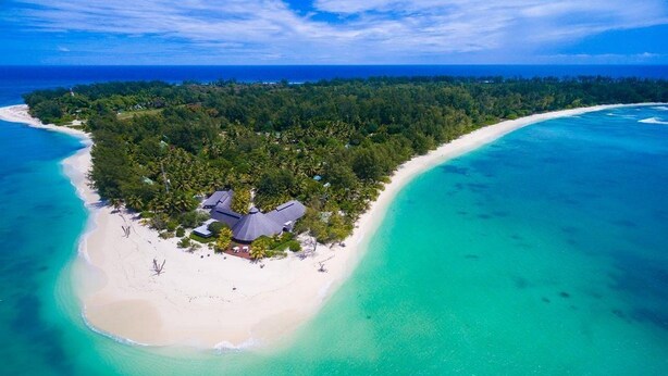 Vista aérea de uma ilha tropical com praia, floresta densa e um pequeno edifício com telhado de palha.