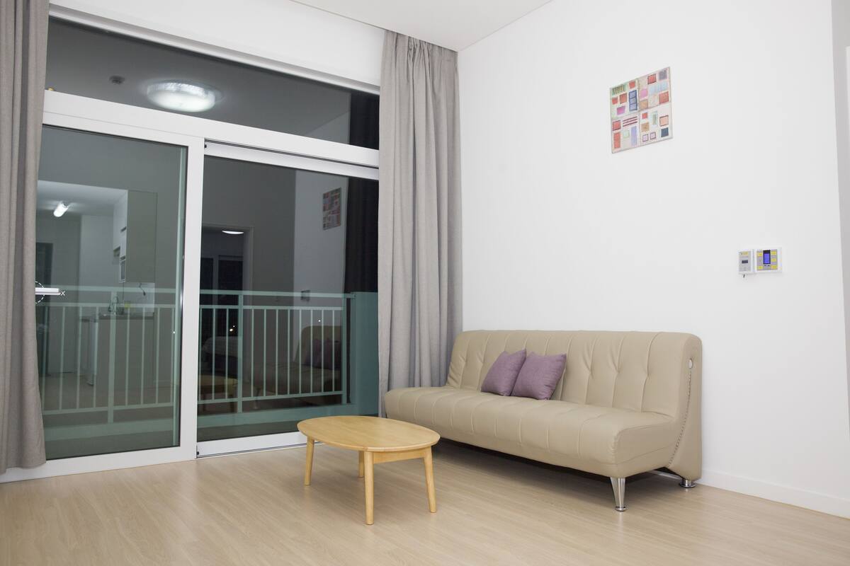 A modern living room with a sliding glass door leading to a balcony, a beige sofa, and a small wooden table.