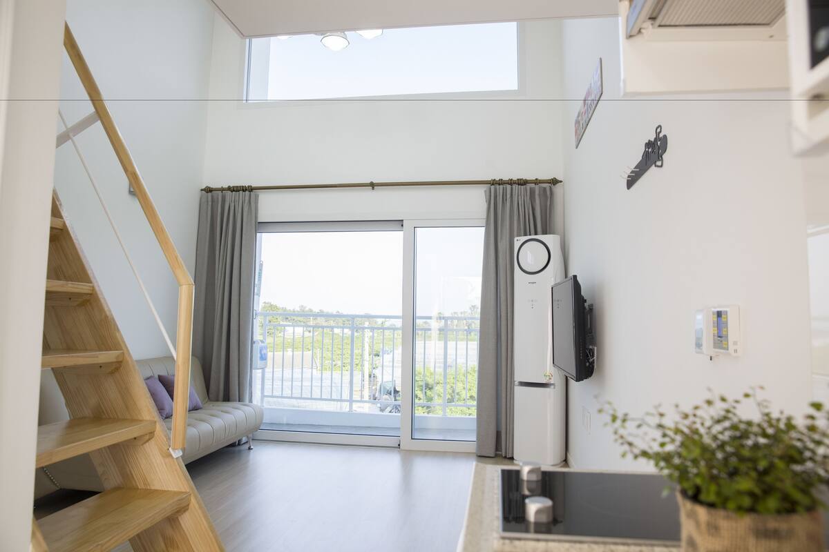 A modern apartment with a wooden staircase, a sliding glass door to a balcony, a built-in air conditioning unit, and a small kitchen area.
