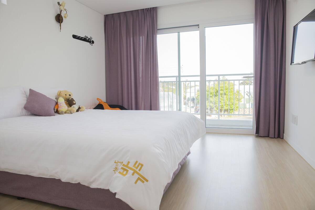 A bedroom with a large bed, a balcony with curtains, and a view of the outside.