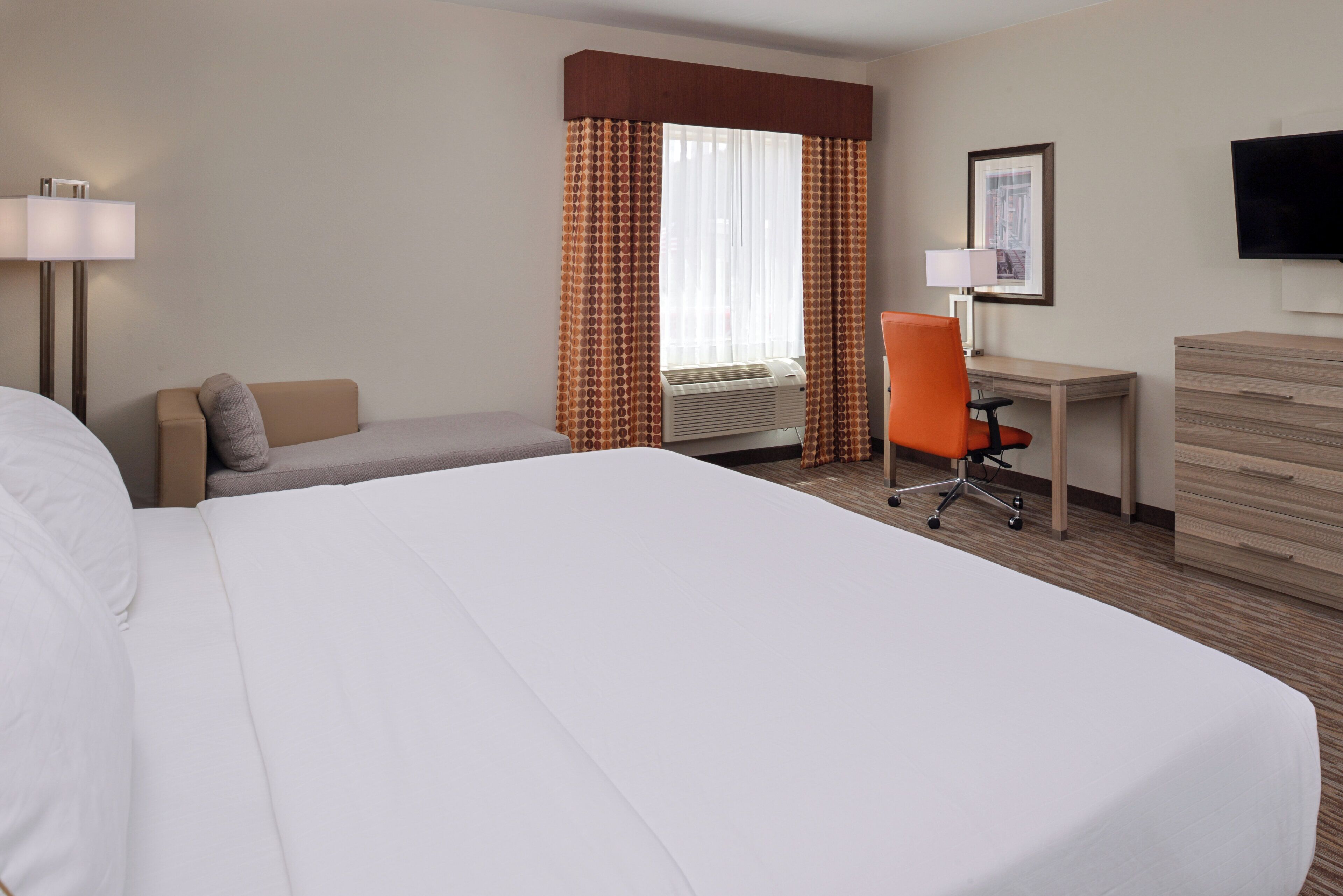 Une chambre d’hôtel avec un grand lit, un bureau, une chaise de bureau, une commode, une télévision et une fenêtre avec des rideaux.