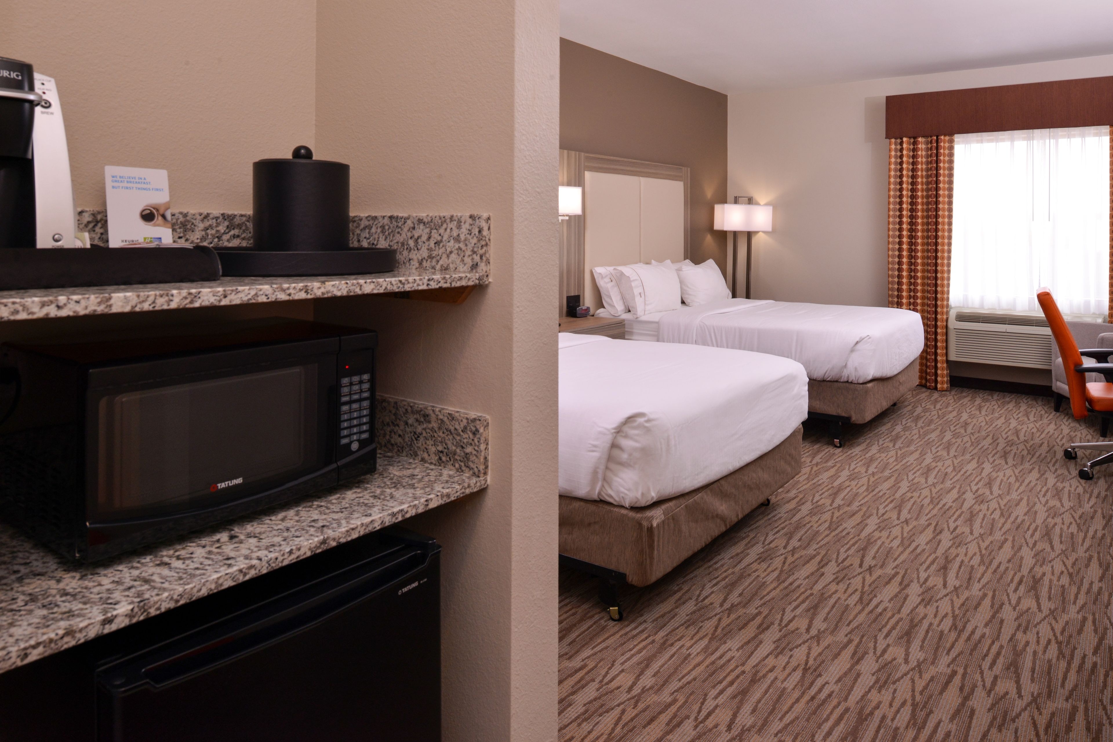 A hotel room with a kitchenette, two beds, a desk, and a window with curtains.