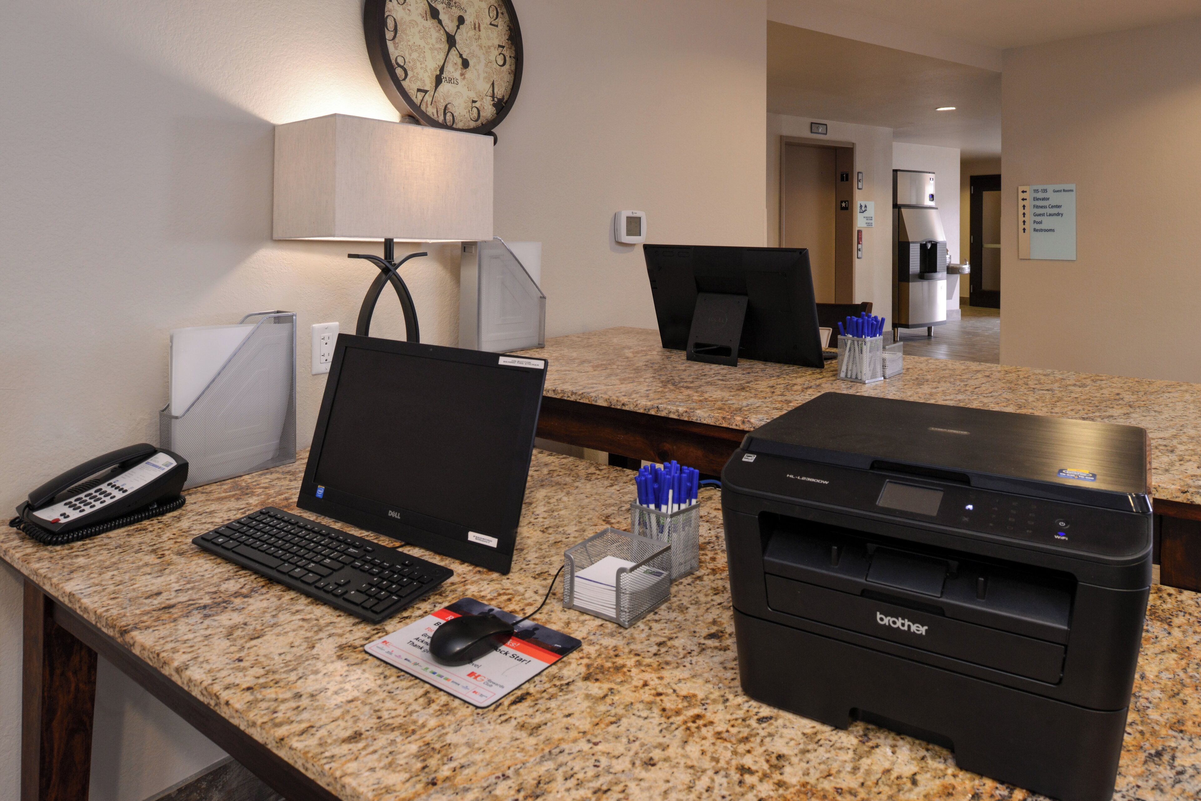 A hotel reception desk with a desk phone, computer, printer, and a clock on the wall.