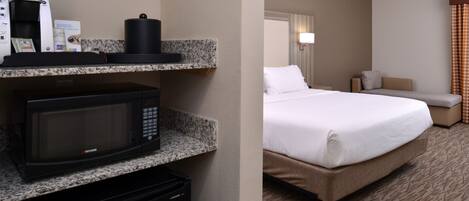 A hotel room with a kitchenette area including a microwave, coffee maker, and a small refrigerator. There is a bed with a white comforter and a bedside table with a lamp.