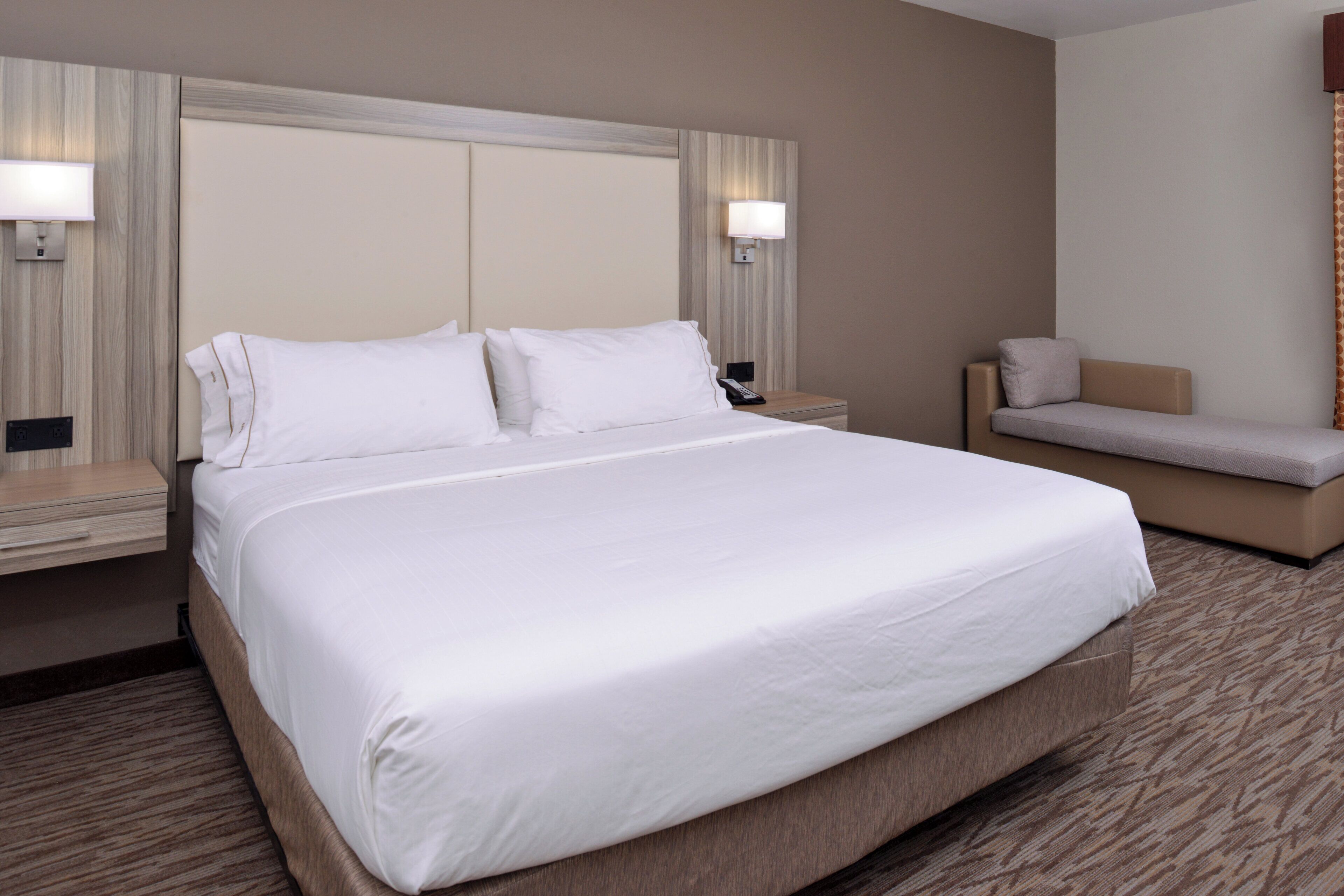 Habitación de hotel con una cama grande, mesitas de noche, un sofá y lámparas fijadas a la pared.