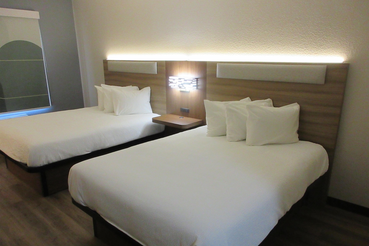 A hotel room with two beds, a nightstand, and a wall-mounted lamp.