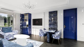 A modern living room with a round white coffee table, blue patterned sofas, and matching chairs. There is a dining area with a marble table and chairs, a chandelier, and a TV mounted on the wall.