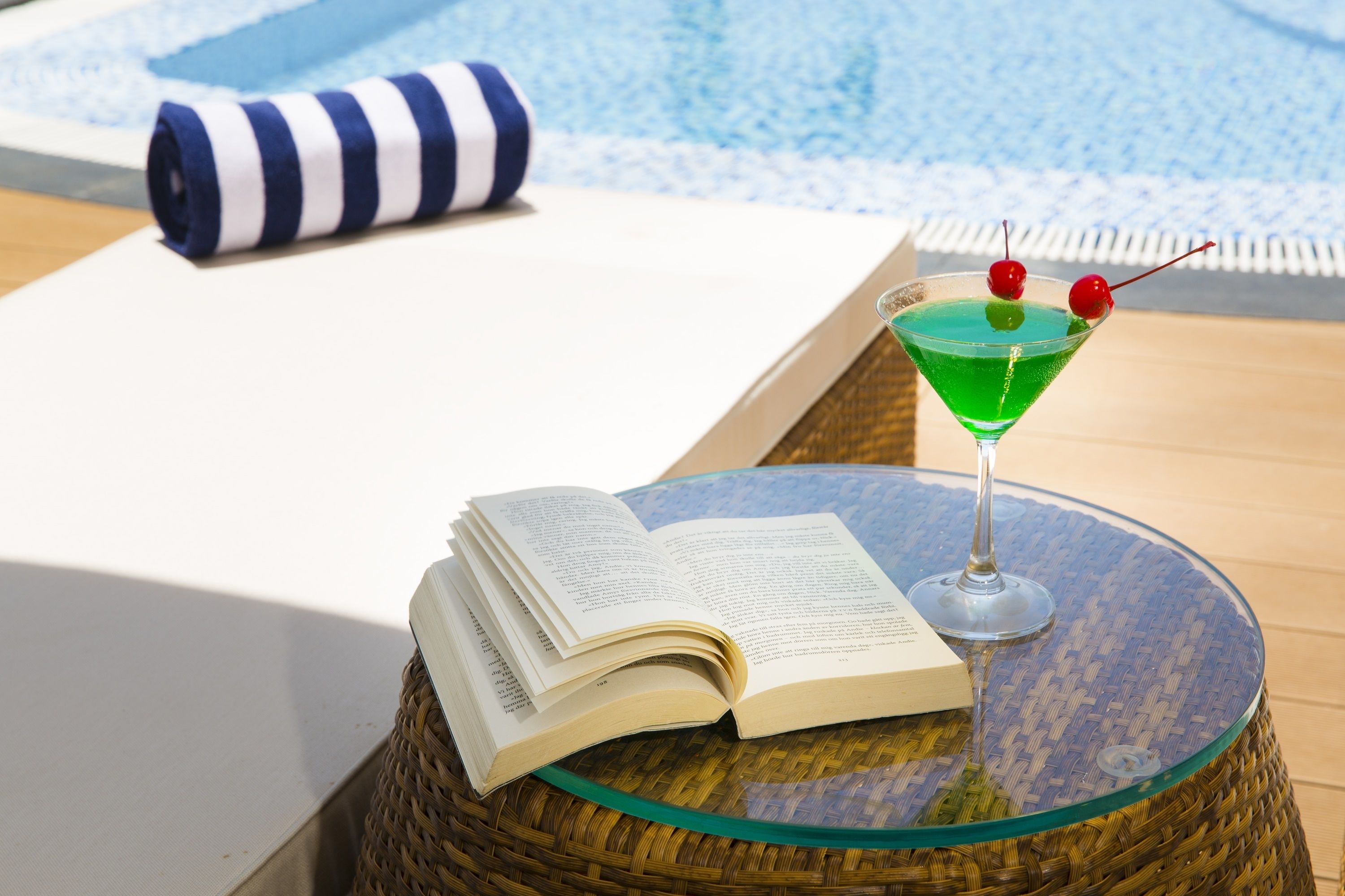A book on a glass table, a striped towel, and a green drink with cherries.