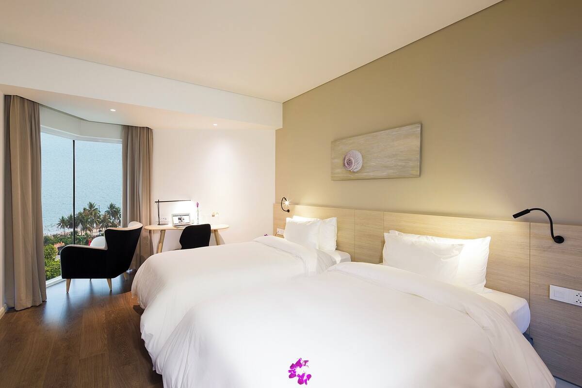 A hotel room with two beds, a desk, a chair, and a large window with a view of palm trees.