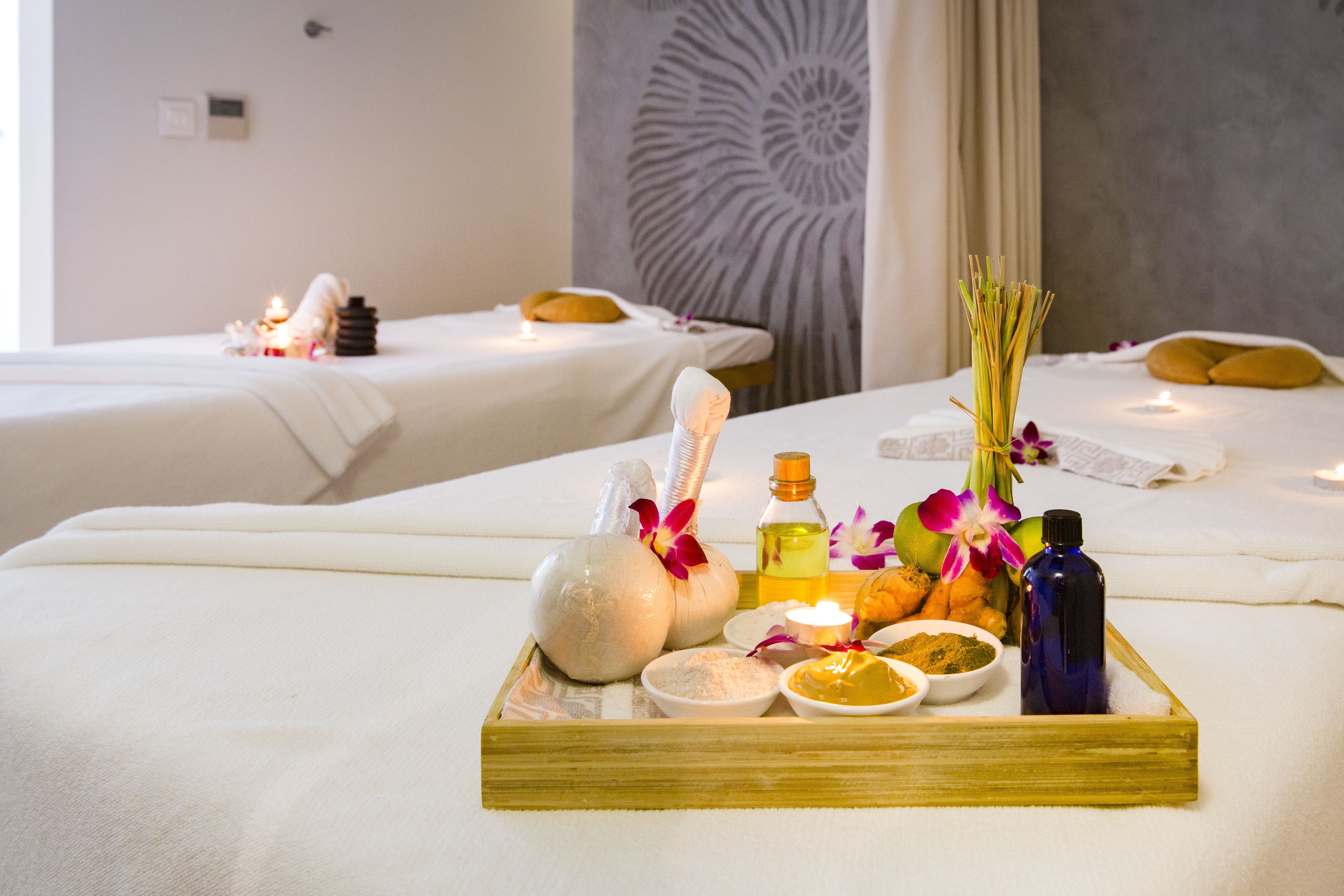 A spa room with two beds, each with a towel and a candle. A tray with bath products and flowers is on the bed.