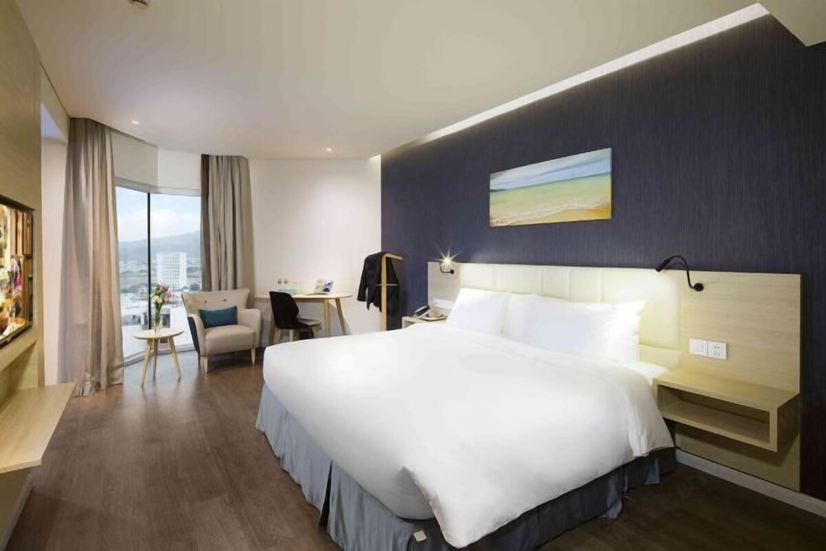 A modern hotel room with a large bed, a desk, a chair, and a view of the city through large windows.