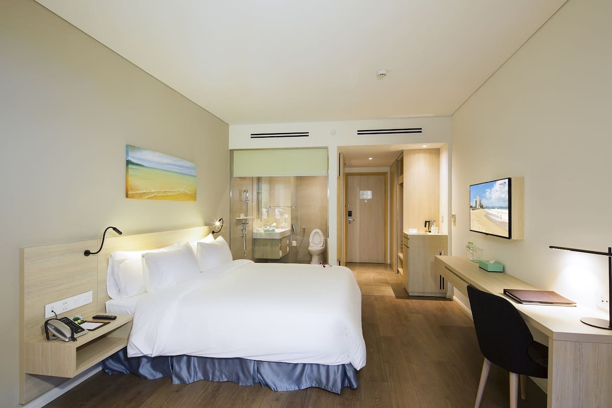 A hotel room with a large bed, bedside table, desk, and chair. There is a bathroom with a sink and mirror.
