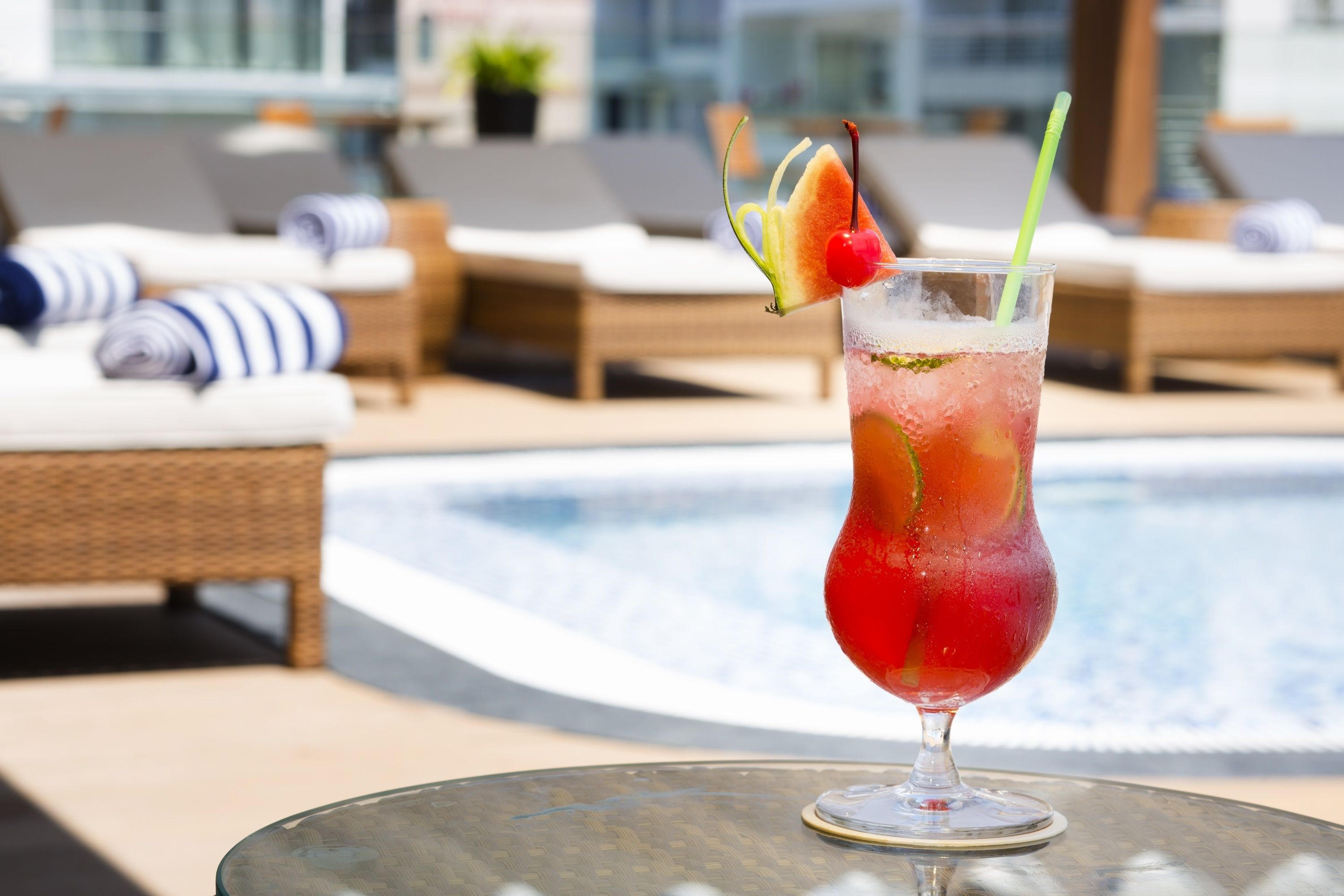 A glass of red cocktail with a cherry and a slice of fruit on a glass table by a poolside lounge area.