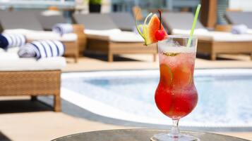 A glass of red cocktail with a cherry and a slice of fruit on a glass table by a poolside lounge area.