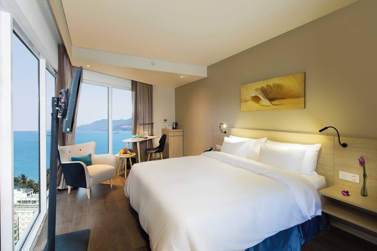 A hotel room with a large bed, a desk, a chair, and a view of the sea.