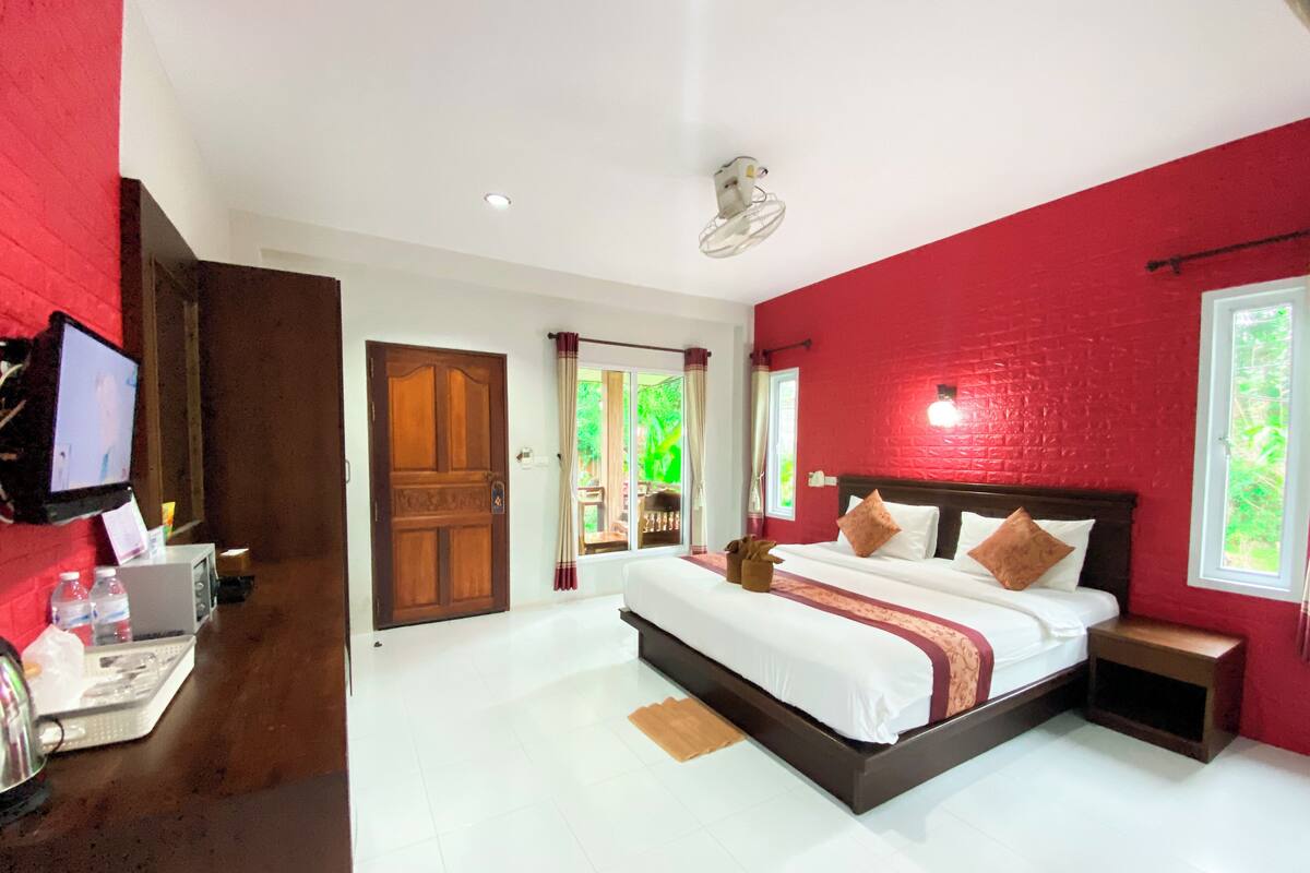 A hotel room with a large bed, a television, a desk with a computer, and a window with a view of greenery.