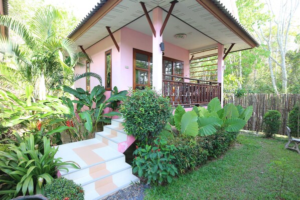 A pink building with a covered porch, surrounded by lush greenery and a wooden fence.