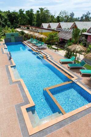 A long swimming pool with lounge chairs and thatched umbrellas, surrounded by palm trees and buildings with red roofs.