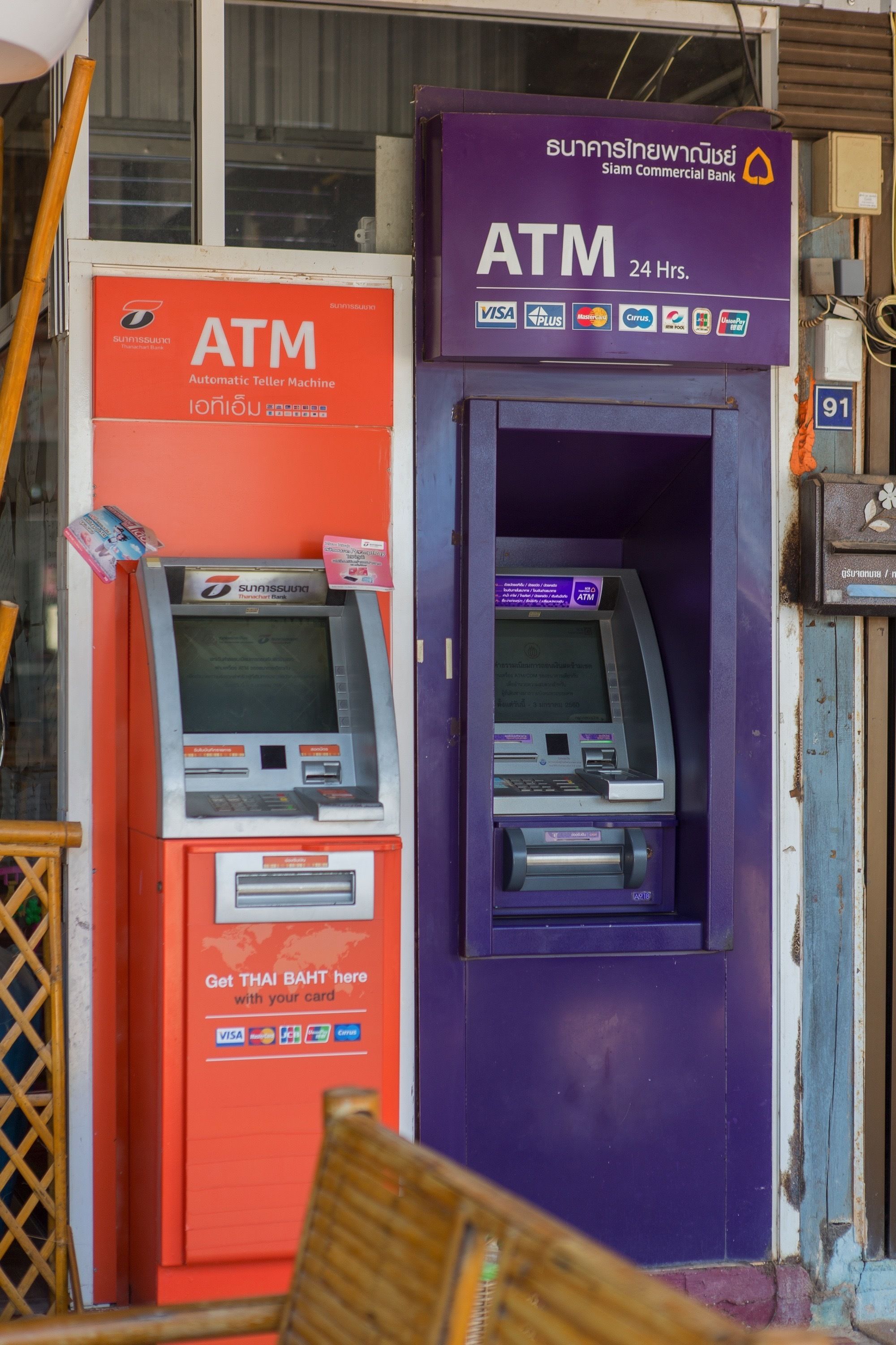 Deux distributeurs automatiques de billets, l’un rouge et l’autre violet, avec une pancarte au-dessus du distributeur violet indiquant « Distributeur ouvert 24 h/24 ».
