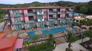 Un hotel de varios pisos con zona de piscina, balcones y abundante vegetación al fondo.