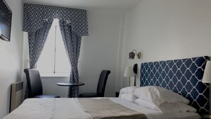 Ein Hotelzimmer mit einem Bett, zwei Stühlen, einem kleinen Tisch, einer Lampe und gemusterten Vorhängen.