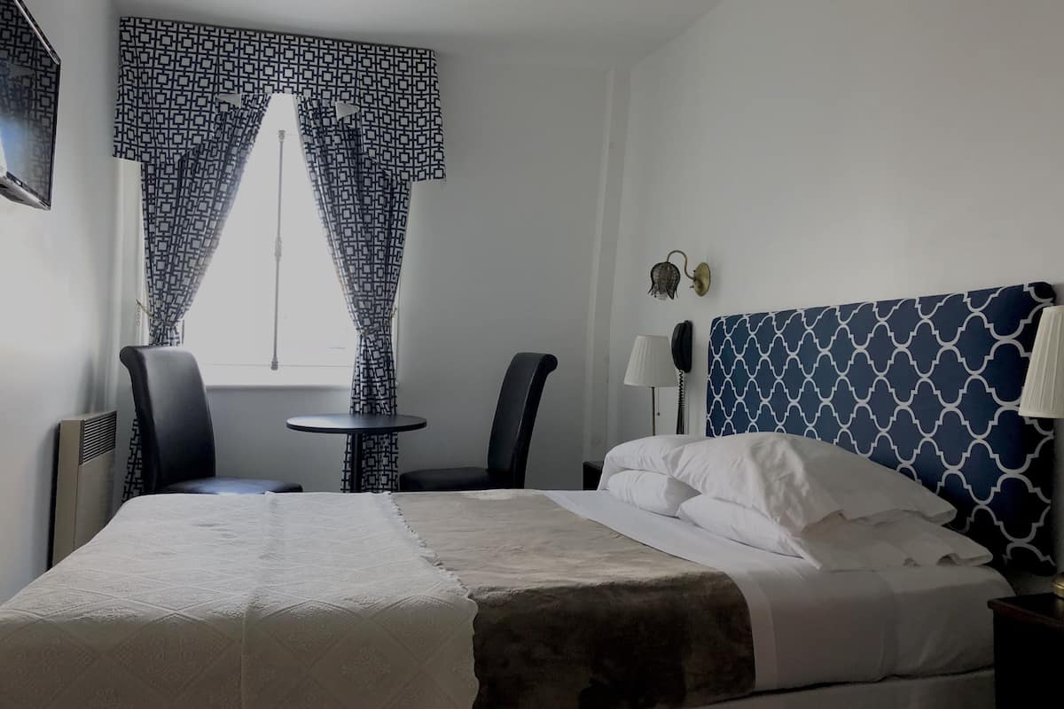 Hotellihuone, jossa on sänky, kaksi tuolia, pieni pöytä, lamppu ja kuviolliset verhot.
