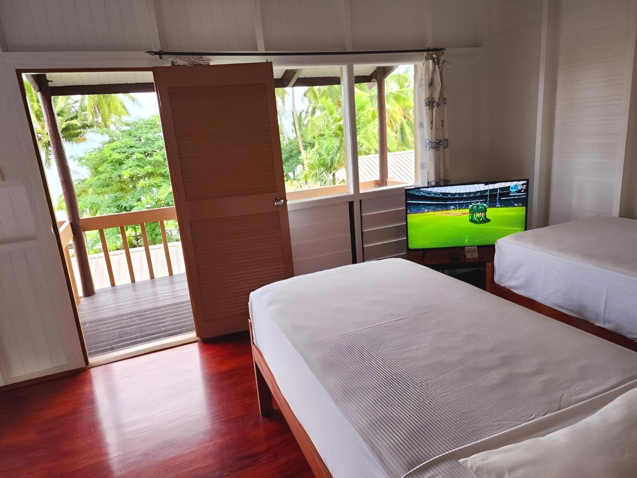 Habitación con dos camas, un televisor sobre un mueble bajo, piso de madera y puerta corredera que da a un balcón con vistas a vegetación.