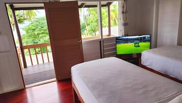 Habitación con dos camas, un televisor sobre un mueble bajo, piso de madera y puerta corredera que da a un balcón con vistas a vegetación.